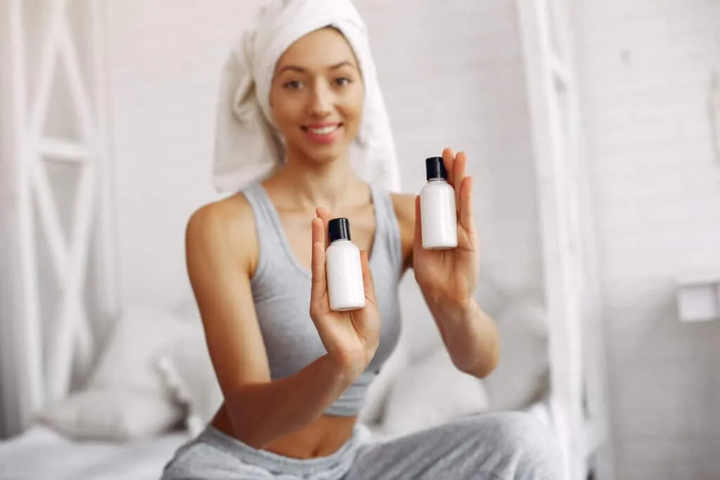 Girl in a studio. Lady with cream. Woman in a gray t-shirt. Girl sitting on a bed. mini plastic bottles