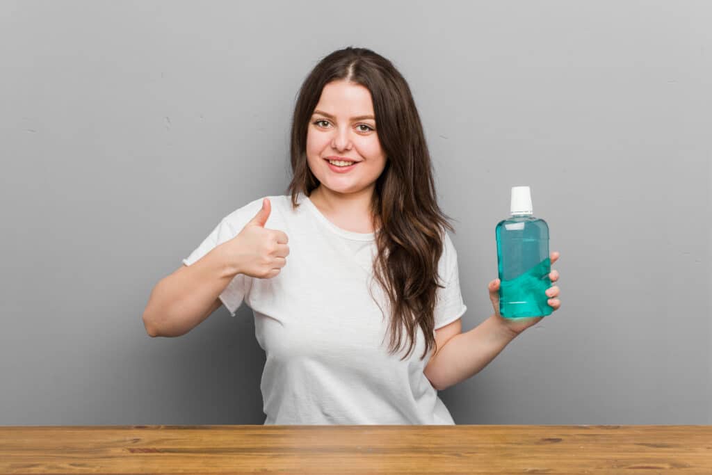 Young plus size curvy woman holding a mouthwash made of 16 oz plastic bottle smiling and raising thumb up