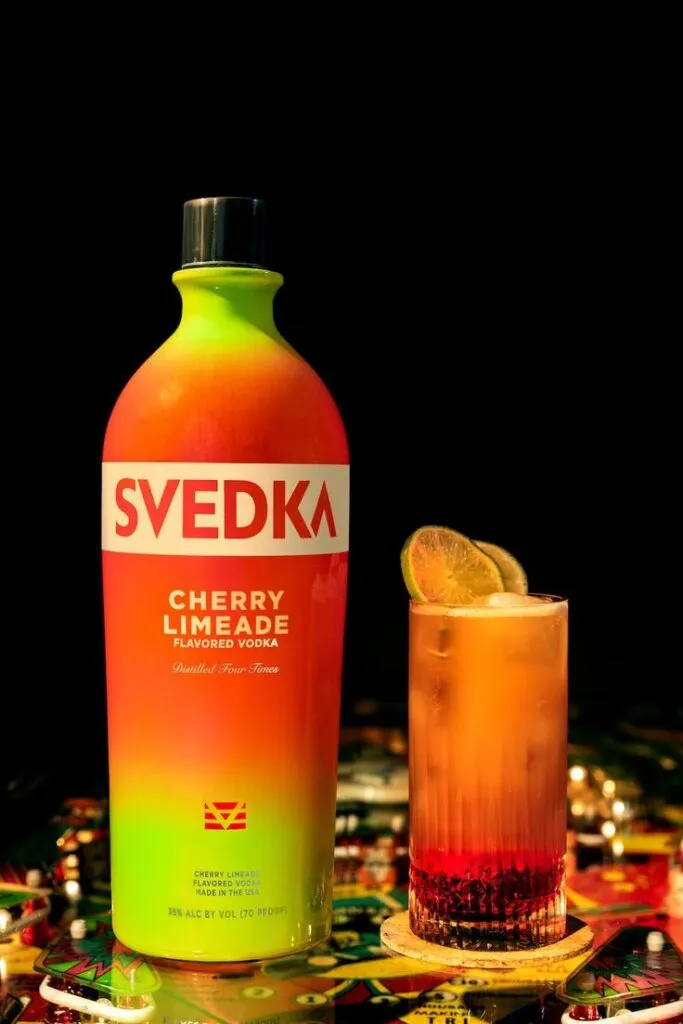 A bottle of Svedka Cherry Limeade Flavored Vodka in plastic bottle and a glass of the cocktail on a colorful surface.