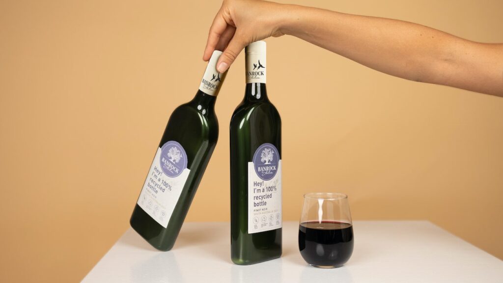 A hand holding two recycled wine bottles , showing the trend of plastic bottles for liquor with a glass of red wine beside them.