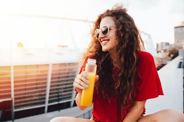 A smiling woman looking away holding a plastic juice container with fresh orange juice. 