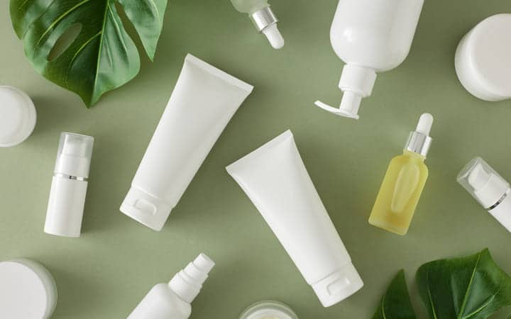 Flat lay of various white cosmetic packaging - tubes, jars, bottles, droppers with green tropical leaves on a calming green backdrop.