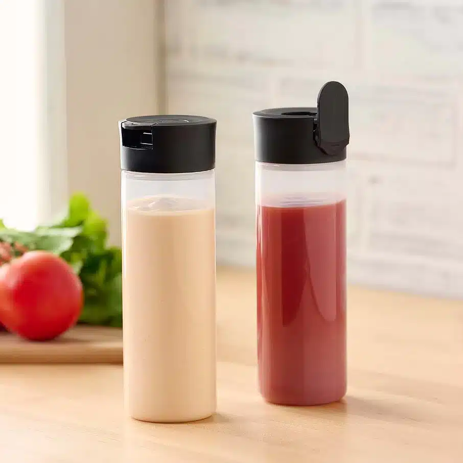 Two clear plastic squeeze bottles with black lids, filled with juices on a kitchen counter. 